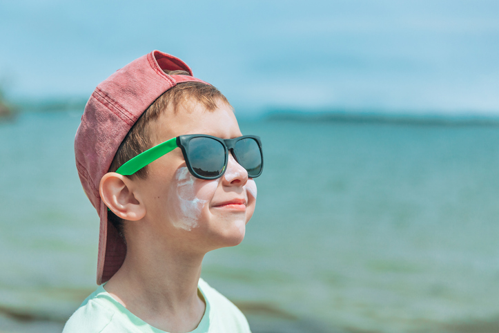 Young boy with sunscreen on his face, wearing sunglasses and a red cap, looking towards the sun at the beach.